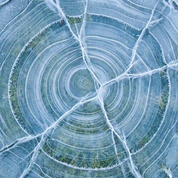 Detailed overhead view of concentric ice rings and radial fractures in frozen pond winter background.