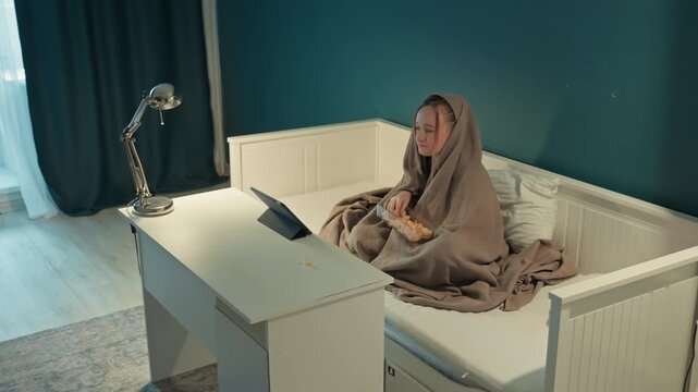Young woman sits on white daybed covered with brown blanket. Woman remains seated as woman interacts. Scene takes place in room with teal walls dark curtains light
