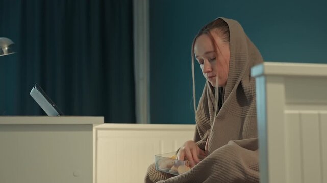 Young woman sits on bed wrapped in brown textured blanket. Woman holds and eats from clear plastic container. Room features teal walls dark curtains light colored furniture