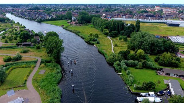 Aerial view video clip of scenic canal river, small boats cruising, skate park, green parkland, waterfront houses, and distant city skyline in Netherlands