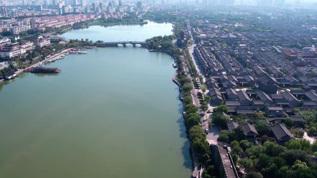 Aerial perspective of a historic town beside a wide waterway, with a stone arch bridge connecting areas, and modern city skyline in distance.