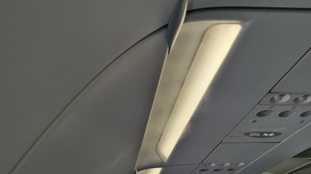 The airplane cabin ceiling presents a long illuminated overhead panel with air vents, reading lights, and passenger service controls. The compact arrangement reflects standard passenger amenities.