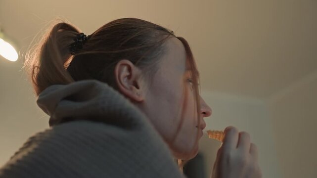 Close up of young woman with brown hair in ponytail wearing earrings. Woman consistently holds food near her mouth. Background features neutral colored walls and soft illumination