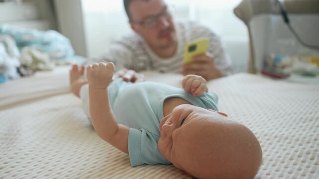Unplugged young father distracted by his smartphone, ignoring his newborn son lying on the bed, highlighting the problem of parental inattention and modern technology addiction