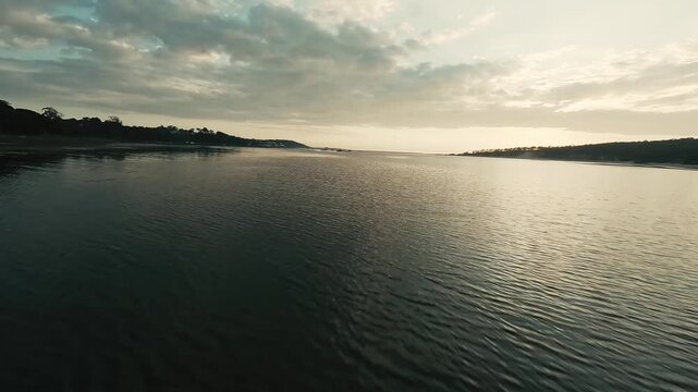 Aerial view of calm water reflecting golden sunset light. Trees line both shores under cloudy sky. Serene and peaceful scene ideal for travel, nature, or meditation projects