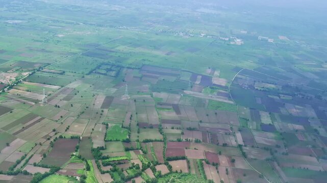 The aerial view from an airplane shows a mosaic of green and brown agricultural fields divided by narrow roads. Tree-lined boundaries and scattered rural settlements form a varied countryside pattern.