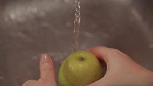 Close up of hands washing green apple under running water in sink. Fruit is rotated and rinsed thoroughly with clear flowing stream. Sink drain visible throughout sequence