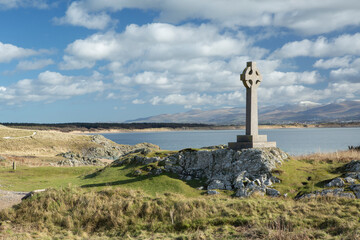 A solitary Celtic cross memorial stands on rocky grassland above a calm sea, framed by rolling dunes, distant mountains and a bright sky with scattered clouds,  © © Raymond Orton