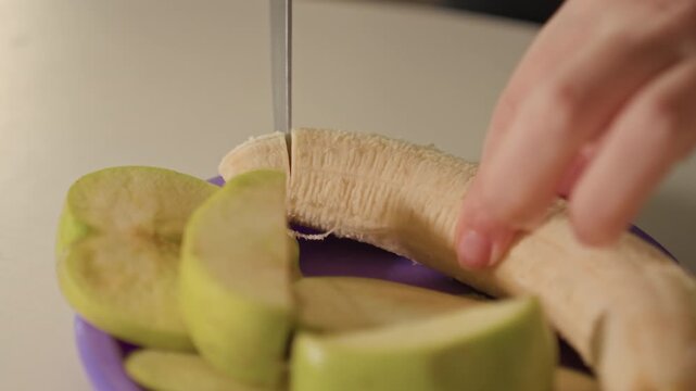 Close up of hand slicing green apples and bananas on purple plates. Knife moves steadily through fruit creating thin slices. Plain background emphasizes focus on food preparation