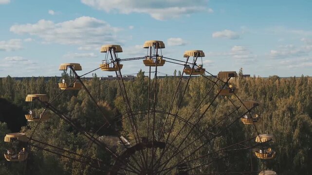 Rusting Ferris wheel arc with yellow scalloped cabins above treeline, weathered spokes, pale blue sky, distant buildings, and quiet amusement park atmosphere at daylight
