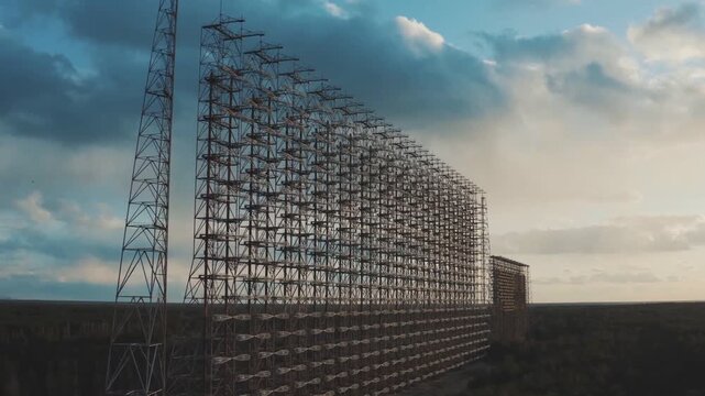 Curved desert antenna lattice towers behind barbed fence at sunset, backlit by dramatic clouds across open horizon, showing remote industrial infrastructure in wide landscape
