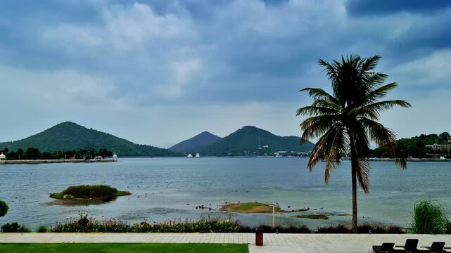 The serene Udaipur lakescape extends toward lush green hills, with a tall coconut palm at the right and small islets across calm blue water. White waterfront structures and a manicured promenade.