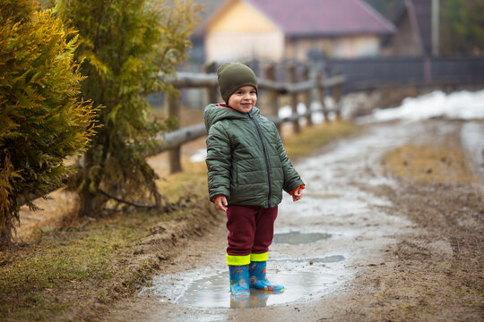 Little boy in protective rubber boots and rain clothes jumping in mud puddle. Happy child having fun while playing in puddle after rain.