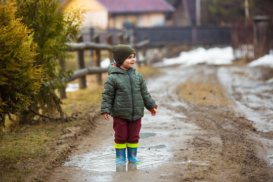 Little boy in protective rubber boots and rain clothes jumping in mud puddle. Happy child having fun while playing in puddle after rain.