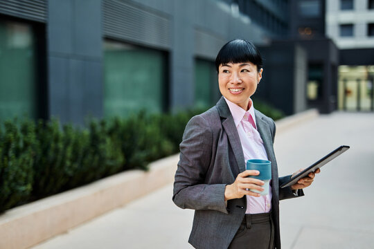 Portrait of a young asian japanese businesswoman woman using a tablet computer and holding a cup of coffee  in a city park, surrounded by modern corporate office buildings