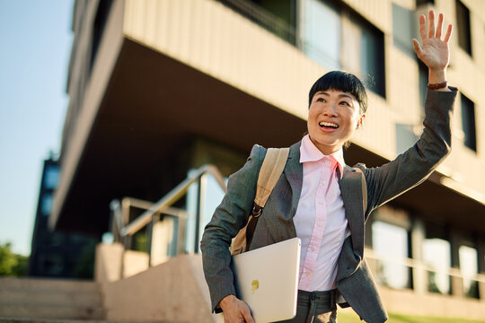 Portrait of a young asian japanese businesswoman woman walking down the street and waiting and waving for ataxi cab, surrounded by modern corporate office buildings