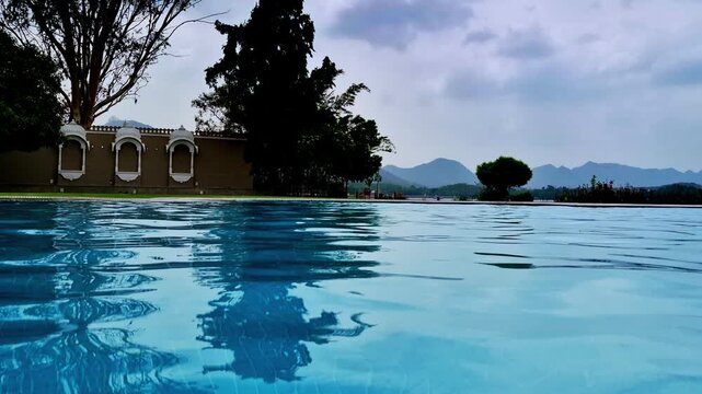 The broad turquoise swimming pool complements the heritage-style ochre building, ornate white arched windows, mature trees, and manicured greenery. Distant rugged mountains frame Udaipur.