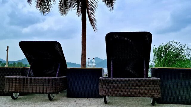 Two woven wicker lounge chairs and bottled water sit beside a tall coconut palm and ornamental grasses. Lush hills and a lake extend beyond the terrace beneath a cloudy blue sky in Udaipur.