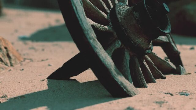 A wagon wheel is resting on the sandy ground close to large rocks. The scene shows remnants of history in an outdoor location. Sunlight creates shadows around the wheel.