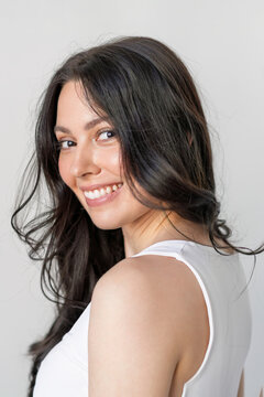 Smiling woman with long dark hair in white tank top posing on neutral background