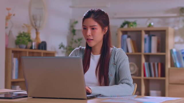 Young Asian woman laptop home office desk computer working indoor, student remote work study workspace with notebook shelf plant focused calm expression