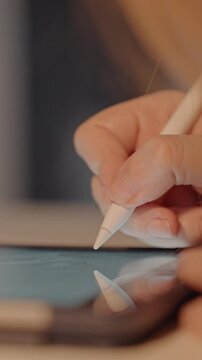 Close up of hand holding white stylus. Fingers grip digital pencils moving them. Blurred indoor background suggests warm lighting and neutral tones focusing on close up details