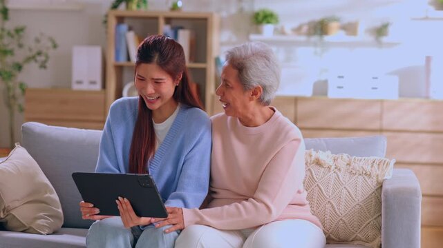 Asian woman elderly using tablet in living room smile with family on couch at home sharing digital device support bonding care cozy interior indoors