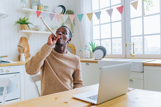 African american man celebrating his birthday virtually at home, wearing a party hat, blowing a party horn while looking at a laptop in a kitchen decorated with bunting