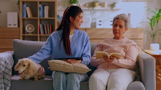 Senior woman dog on sofa reading book with tablet at home living room family caregiving cozy companionship warm indoor lifestyle scene relaxed pet gathered on couch
