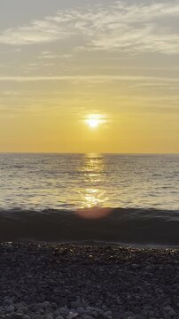 Vertical video of a beautiful golden sunset over the sea, with gentle waves lapping on the pebble beach.