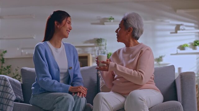 Senior woman young caregiver family living room water smile conversation sofa home care support drink cozy interior health connection warm happy gentle shared moment tidy bright indoor setting