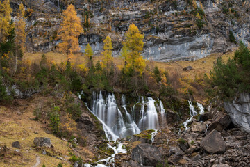 Clear mountain water cascades over rocky terrain beneath golden autumn larches at Siebenbrunnen, Switzerland. © Andrei Tomas
