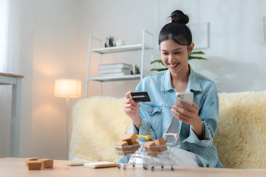 Happy woman shopping online with smartphone and credit card at home, using e-commerce and mobile payment with miniature carts, packages for convenient digital retail delivery.