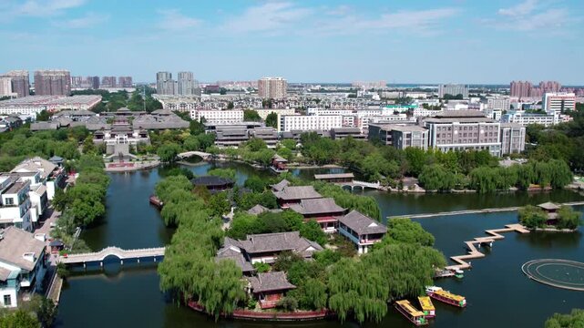 Aerial view of a traditional garden with lakes and pavilions, adjacent to modern urban buildings under a clear blue sky.