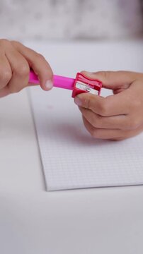 A kid boy around eight years old sharpens a yellow pencil using a red and pink pencil sharpener while sitting at a table in a bright room filled with natural light.