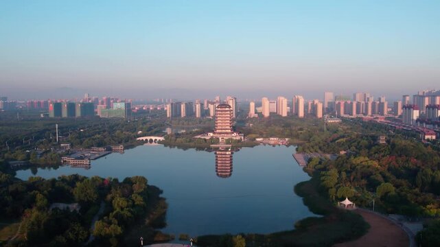 Aerial view of a city park with a lake, a traditional tower, and surrounding urban buildings under clear sky.
