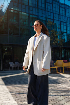 Pretty woman standing in elegant white blazer near contemporary office building. Portrait of female model wearing sunglasses with business style and urban lifestyle concept.