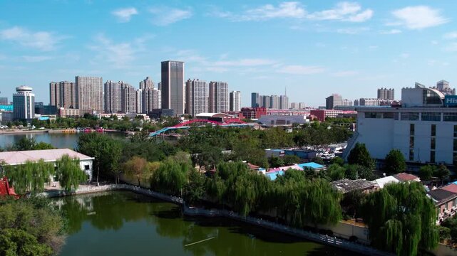 A serene urban park with a lake and traditional pavilion, bordered by dense trees and modern high-rise buildings under a clear blue sky.