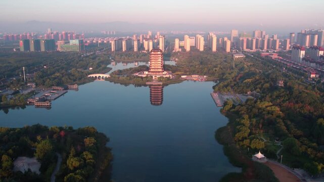 A traditional pagoda stands on an island in a calm lake, with city buildings and green parks around at dawn.