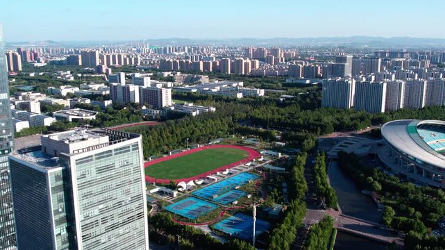 Aerial shot of a modern urban area featuring a sports complex, high-rise buildings, green spaces and a waterway under clear sky.