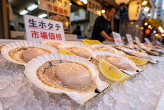 Fresh raw sea scallops in shells served on crushed ice with lemon slices at a bustling Japanese fish market stall with Japanese signage and vendor.
