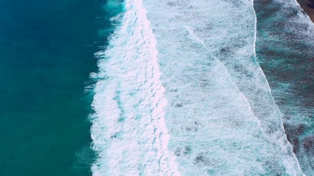 Capture the hypnotic power of the ocean with this mesmerizing top-down aerial shot of powerful waves rolling toward the coast. Showcasing a stunning contrast between deep emerald waters and bright whi