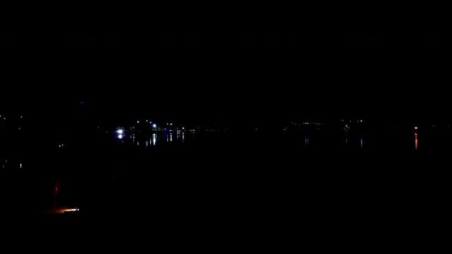 Udaipur's dark lakefront features scattered shoreline lights casting shimmering reflections across calm water. The night setting creates a quiet and subdued waterfront scene.