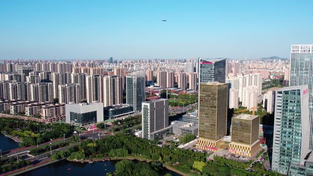 A wide aerial shot of a modern urban area with high-rise buildings, a waterfront, and green spaces under a clear blue sky.
