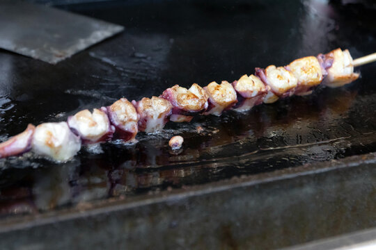 Grilled octopus skewer on hot metal grill with charred edges and glistening oil, savory street food sizzle