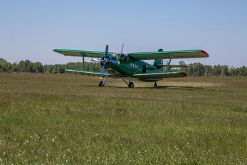 Green Antonov AN-2 biplane aircraft with spinning propeller standing on grass airfield surrounded by trees under clear blue sky, aviation transportation scene © seregraff