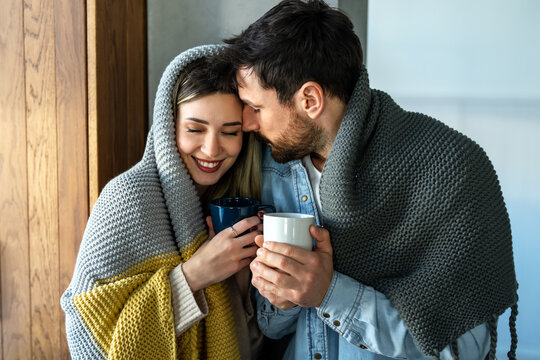Happy young couple with hot drinks cuddling under warm blanket at home