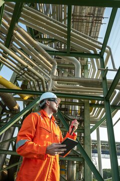 Inspector technician inspecting petroleum engineering in oil refinery performance checklist inspection at fuel industrial plant Male worker holding tablet and radio near oil pipeline structure
