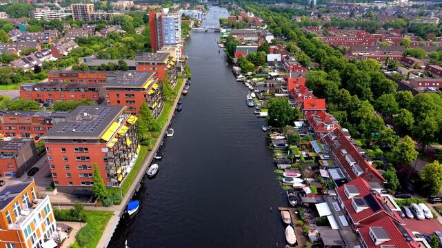 High angle aerial drone video clip of urban river canal, modern apartments with solar panels, waterfront houses, small boats moored at docks, car parking lot, and city skyline in Netherlands, Europe