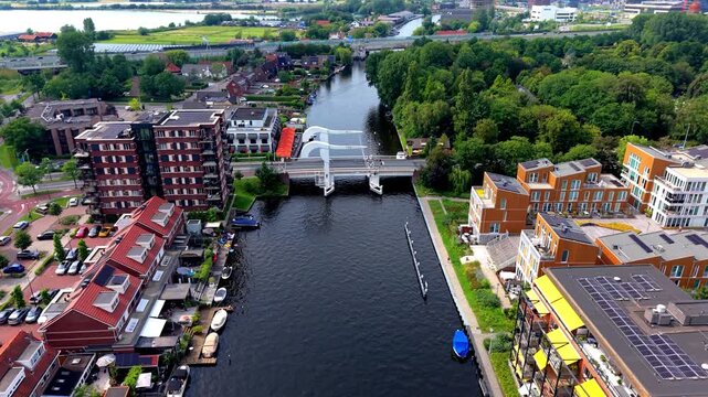 High angle aerial drone video clip of white drawbridge spanning urban river canal, modern apartments with solar panels, waterfront houses, small boats, car parking lot, and lake in Netherlands, Europe